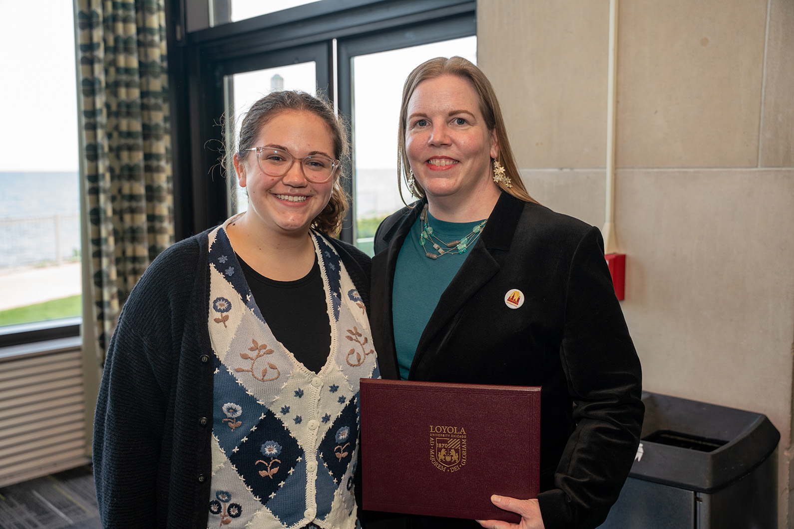 blonde woman in teal holds award and poses with curly haired brunette student with glasses